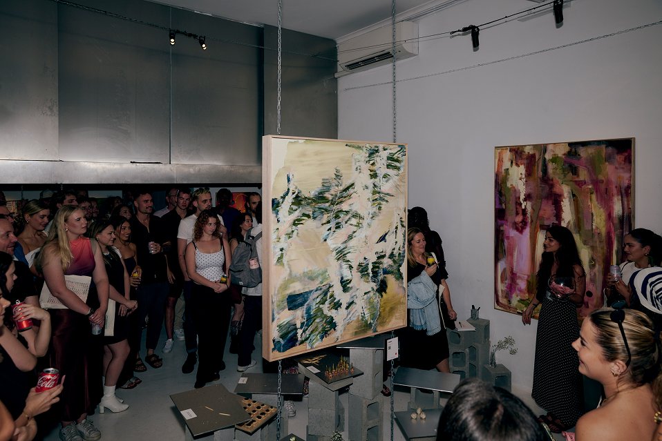 Crowded art exhibition event with overhead track lighting highlighting large paintings and guests